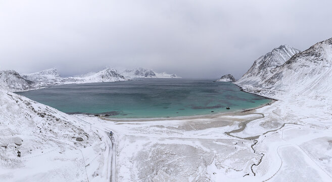 Aerial view of turquoise waters meet snow-laden shores amidst rugged mountains, a serene tableau of nature's contrasting beauty, Nordland, Norway.