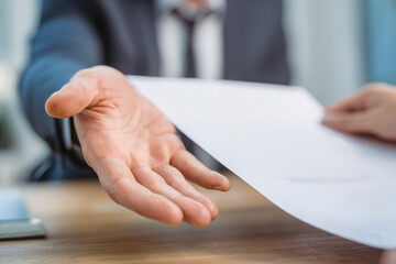 Obraz premium A businessman wea a suit extends a blank white document across a wooden desk offe it to another person for consideration and review.