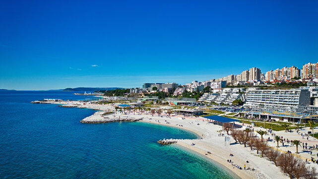Aerial view of sun-kissed beaches meet the azure Adriatic Sea, framed by modern buildings and rocky coastline, Split, Split-Dalmatia County, Croatia.