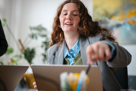 A young woman in a blazer and collared shirt works on a tablet at a table, engaged in a collaborative meeting with colleagues.