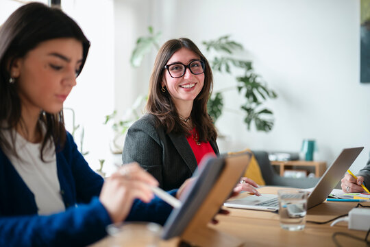 Two women collaborate at a table, one using a tablet and stylus, the other typing on a laptop, with a third person in the background.