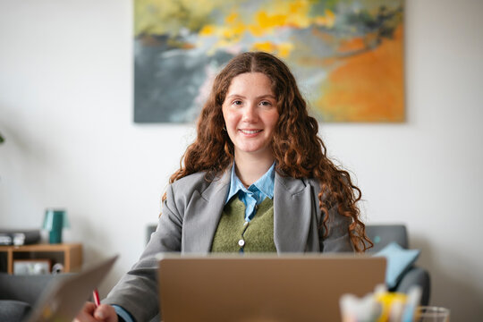 A young woman with curly brown hair smiles while working on her laptop in a bright office setting.