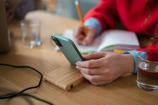 A person is writing in a notebook while holding a smartphone on a wooden desk.