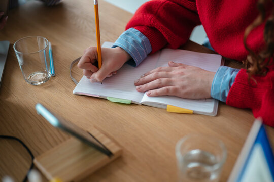 A person's hands are writing in a notebook with a pencil, surrounded by study materials on a wooden desk.