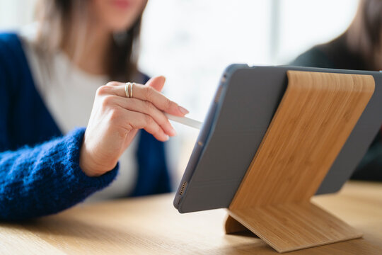 A woman's hand, adorned with rings, uses a stylus to interact with a tablet device resting on a wooden stand.