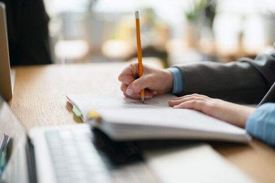 A person in a suit jacket and blue shirt writes with a pencil in a notebook on a wooden desk, with a laptop nearby.