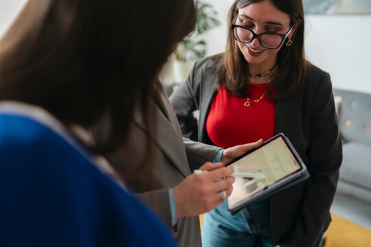 Two women collaborate on a digital tablet, one pointing with a stylus, discussing content on the screen in a modern office setting.