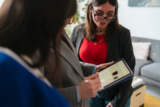 Two women collaborate on a tablet, reviewing product images and data, suggesting a business meeting or shopping decision.