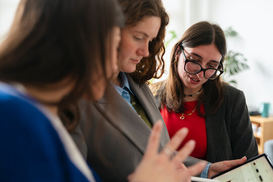Three women in business attire collaborate around a tablet, discussing data and projects in a modern office setting.
