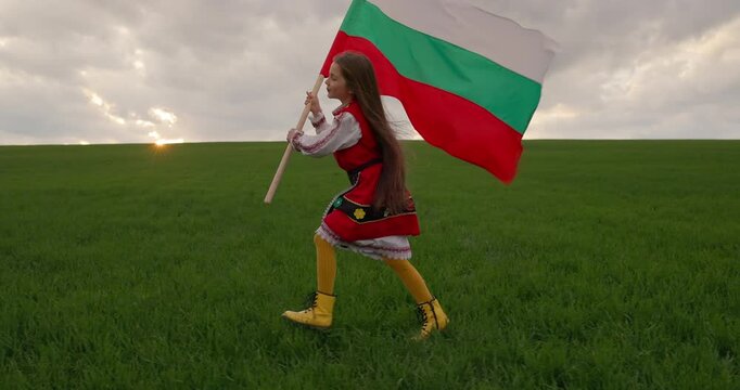 Child in ethnic folklore costume waving Bulgarian flag and run on sunset green agricultural field, bulgarian independence and liberation