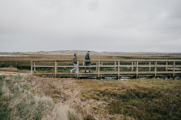 Fototapeta premium A couple is walking across a bridge in a field. The sky is cloudy and the grass is dry
