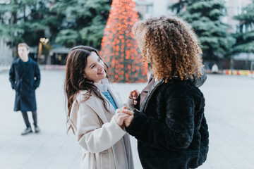 Fototapeta premium Two women hold hands and share a joyful moment in a festive city square with a glowing Christmas tree. The scene shows friends celebrating together as a man watches in the background.