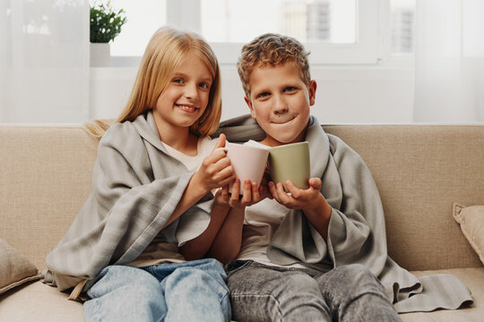 Happy children wrapped in blankets sit on a sofa holding ceramic mugs with warm drinks. Cozy home atmosphere with natural light in the background. Concept of comfort and family time.