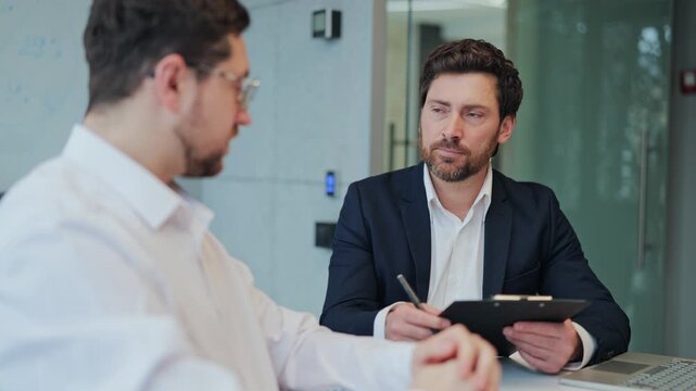 Professionals engaged in a focused business meeting. One takes notes on a clipboard, conversing with a colleague. Serious engagement and collaboration are evident.