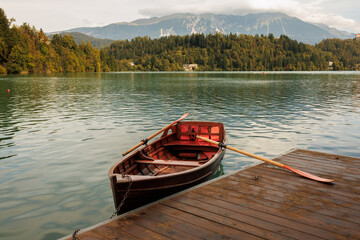Obraz premium A rustic wooden rowboat moored to a dock on a calm lake, surrounded by autumn forest and distant mountains. Stock photo