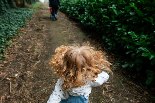 A red-haired, curly-haired girl running free on a path behind her mother, who is pushing the wheelbarrow