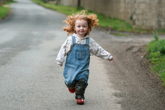 Happy little girl running on the road with red hair flying around her face, muddy dungarees and red boots