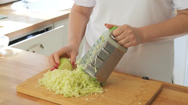 Woman is cutting up a head of cabbage on a wooden cutting board. She is using a grater to shred the cabbage
