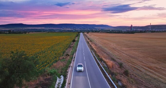 Aerial View of Fast Sports Car Driving Along Country Road Between Sunflower Field and Wheat at Sunset
