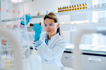 Female scientist wearing protective eyewear and gloves, pouring liquid from a test tube into a beaker during a chemistry experiment © we.bond.creations