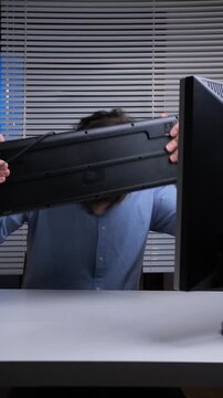 Vertical video stressed employee with an angry facial expression holding a computer keyboard, attempting to smash it against his desktop monitor in a fit of rage at the office
