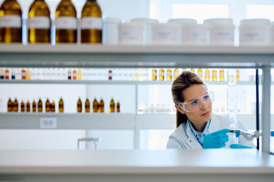 Female scientist wearing protective eyewear and gloves carefully examining a glass condenser, performing scientific research in a laboratory