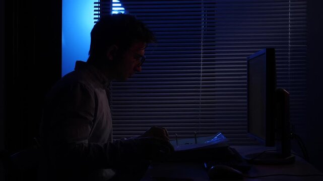 Man in an office working late at night, concentrating on documents under the monitor light, managing paperwork and researching information in a dark atmosphere