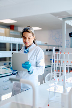 Smiling female scientist wearing lab coat, safety glasses, and gloves, reviewing data on a tablet in a modern lab