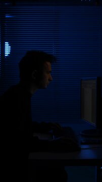 Vertical video man focusing on computer screen, forming a silhouette against a dimly lit window at night, symbolizing cybersecurity, data protection, and a mysterious online presence