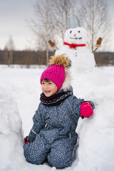 Fototapeta premium A girl in a winter jumpsuit and a pink hat walks in a snowy field in winter, builds a snowman and plays with a dog
