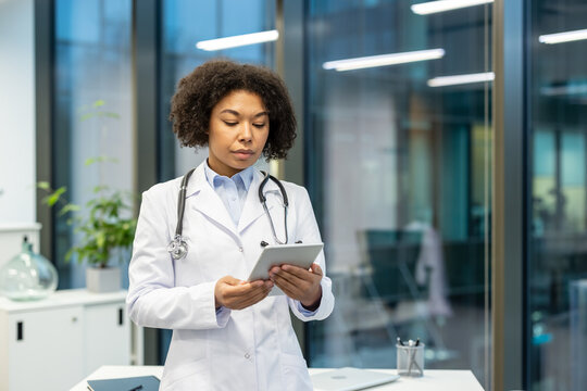 Serious and concentrated young African American female doctor standing in clinic office and using planchet