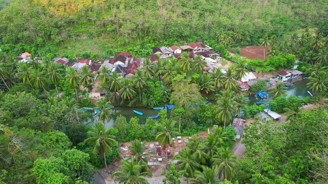 Aerial drone footage of a hidden local village in the middle of a vast forest and mountains, with Maron river and blue touristic wooden boats, in Pacitan regency, Java island, Indonesia