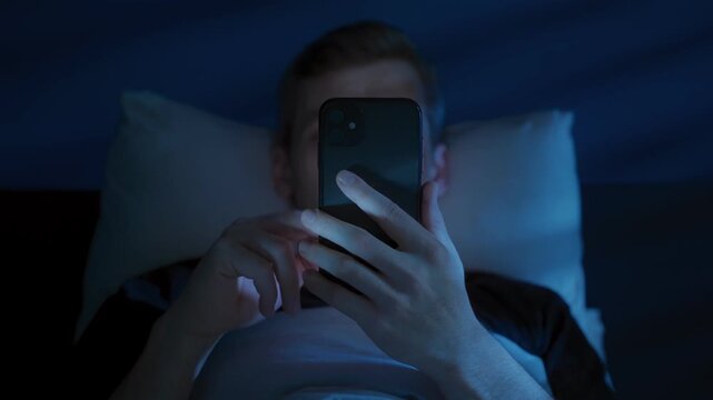Man lies awake in bed late at night, illuminated by the blue light of a smartphone as he endlessly scrolls through social media on mobile phone, struggling with insomnia and digital distraction. 
