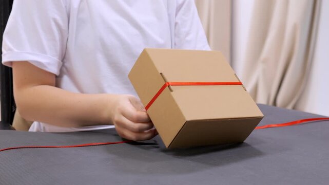 Person is tying a red ribbon around a brown box. The box is on a table. The person is wearing a white shirt