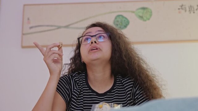 Young woman on hotel bed eating popcorn and reacting to onscreen moment. Glasses reflect TV light as she looks upward and points, expressive face shows surprise and amusement, framed artwork