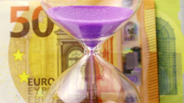 Purple sand dropping through the bulb of the hourglass and euro money on the background. Hourglass with sand stand among money. Deadline, business planning, finance and time is money concept.