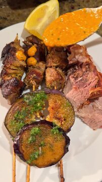 Pouring Mojo Rojo Red Verde Sauce over Grilled Pork Skewers and Roasted Ham Knuckle with Aubergines