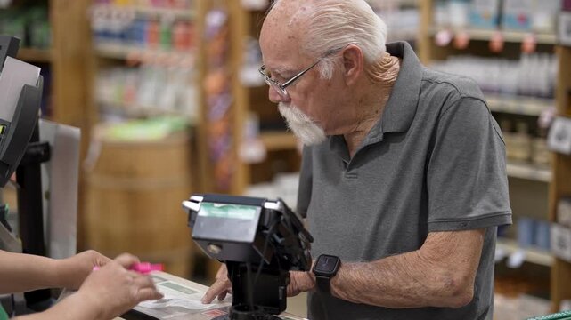 Elderly man scans items at a self checkout in a grocery store and interacts with a staff member for assistance.