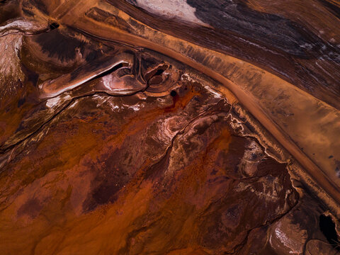Aerial view of swirling, rusty earth tones meeting pale creams and blacks in stark contrast, creating an abstract landscape of textures, Aluminium Refinery, Western Australia, Australia.