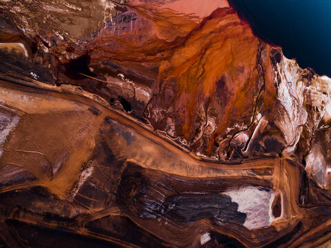 Aerial view of the striated earth unfurls in bands of deep umber and sun-baked ochre, carved by time and industry, Aluminium Refinery, Western Australia, Australia.