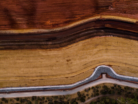 Aerial view of contrasting earth tones meet in an industrial landscape, where rust-red soil gives way to pale fields near a rocky dam, Aluminium Refinery, Western Australia, Australia.