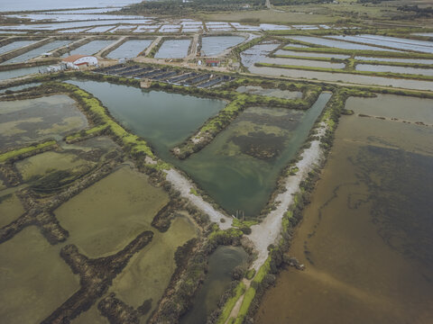 Aerial view of geometric aquaculture ponds reflecting the sky, showcasing the blend of nature and human intervention in the coastal landscape, Moncarapacho, Faro, Portugal.