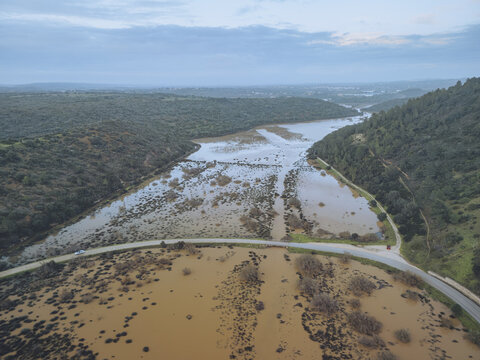 Aerial view of muddy waters stretch through Boca do Rio, reflecting the overcast sky, flanked by verdant hills, Budens, Faro, Portugal.