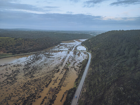 Aerial view of a winding river reflecting the sky, flanked by dense forest and a snaking road, Boca do Rio, Budens, Faro, Portugal.