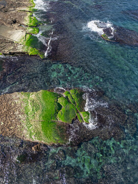 Aerial view of rocky outcrops adorned with vibrant green algae, juxtaposed against the clear turquoise sea, creating a striking contrast of color and texture, Aquaculture, New Taipei City, Taiwan.