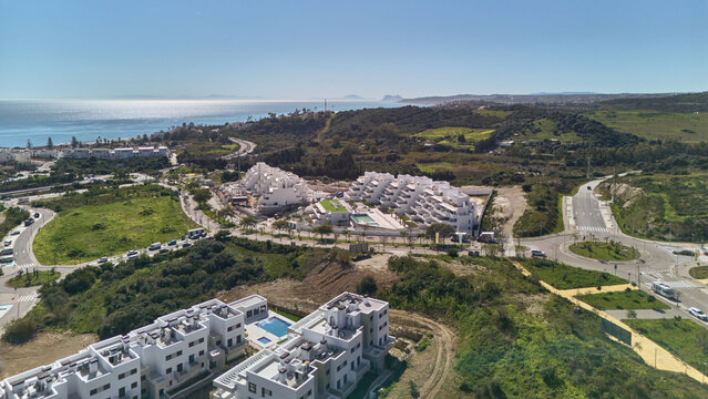 Aerial view of gleaming white residential complexes nestle amidst verdant hills, kissed by the azure sea, creating a striking contrast of urban elegance and natural beauty, Estepona, Andalusia, Spain.
