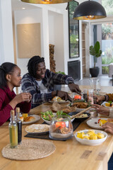 African American friends passing roasted bird around wooden dining table at modern home