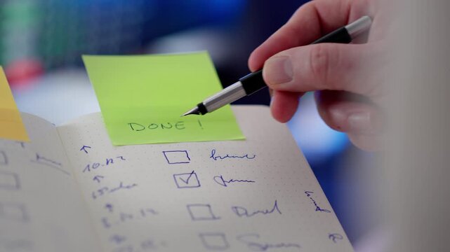 Task checklist notebook DONE sticky note productivity. Closeup hand holding pen marking completed items planner page green reminder paper