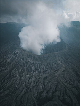 Aerial view of Mount Bromo's volcanic textures rise dramatically, capped with a plume of ethereal smoke against the moody sky, Java, Java, Indonesia.