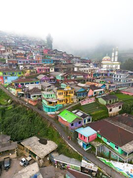 Aerial view of a vibrant village cascading down a hillside, painted in a kaleidoscope of colors, with a mosque standing tall against a misty backdrop, Java, Java, Indonesia.